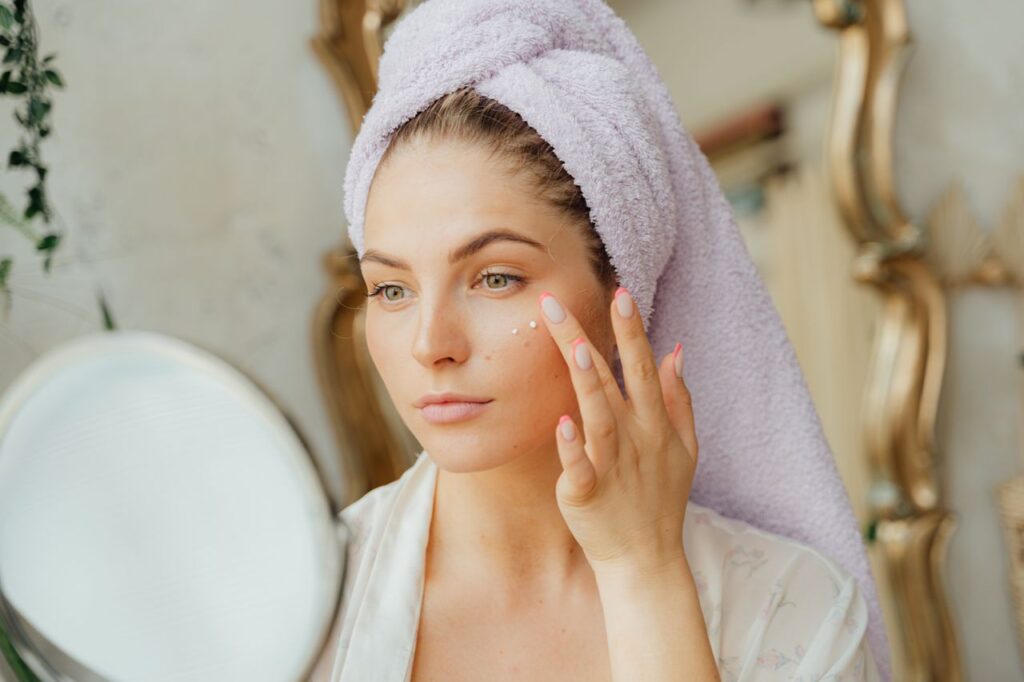 Mulher com toalha na cabeça aplicando creme hidratante no rosto após o banho, cuidado com a pele seca no inverno
